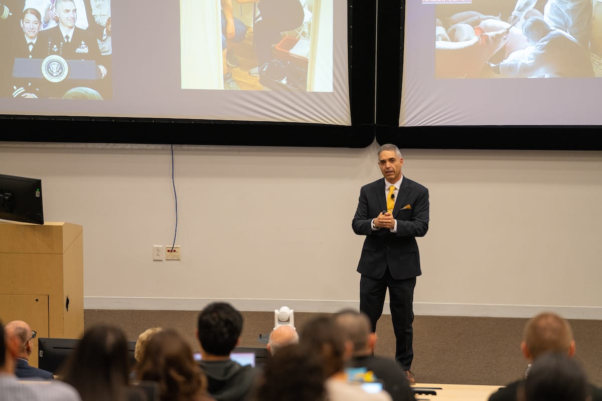 A man in a suit with a yellow tie presenting in front of an audience with slide images projected behind him.