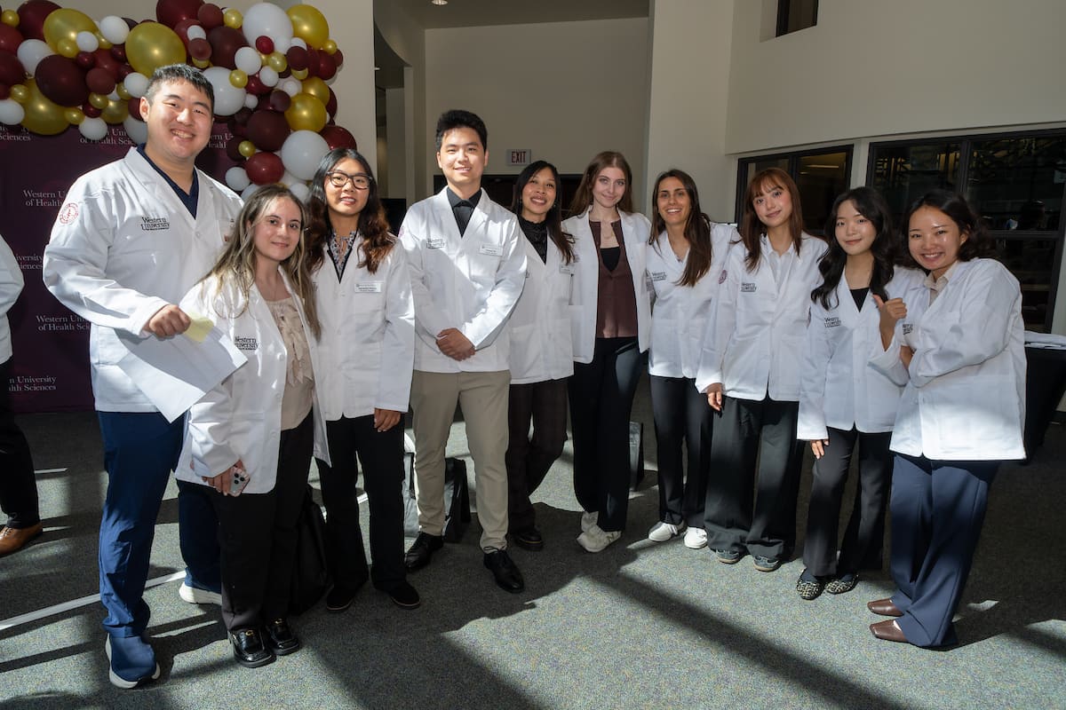 A group of ten people in white lab coats pose indoors, smiling at the camera. There are balloons and sunlight in the background.