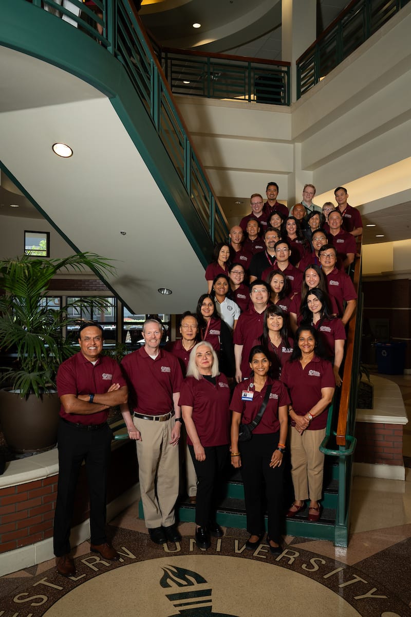 A group of people wearing matching maroon shirts pose together on a staircase in a university building lobby.