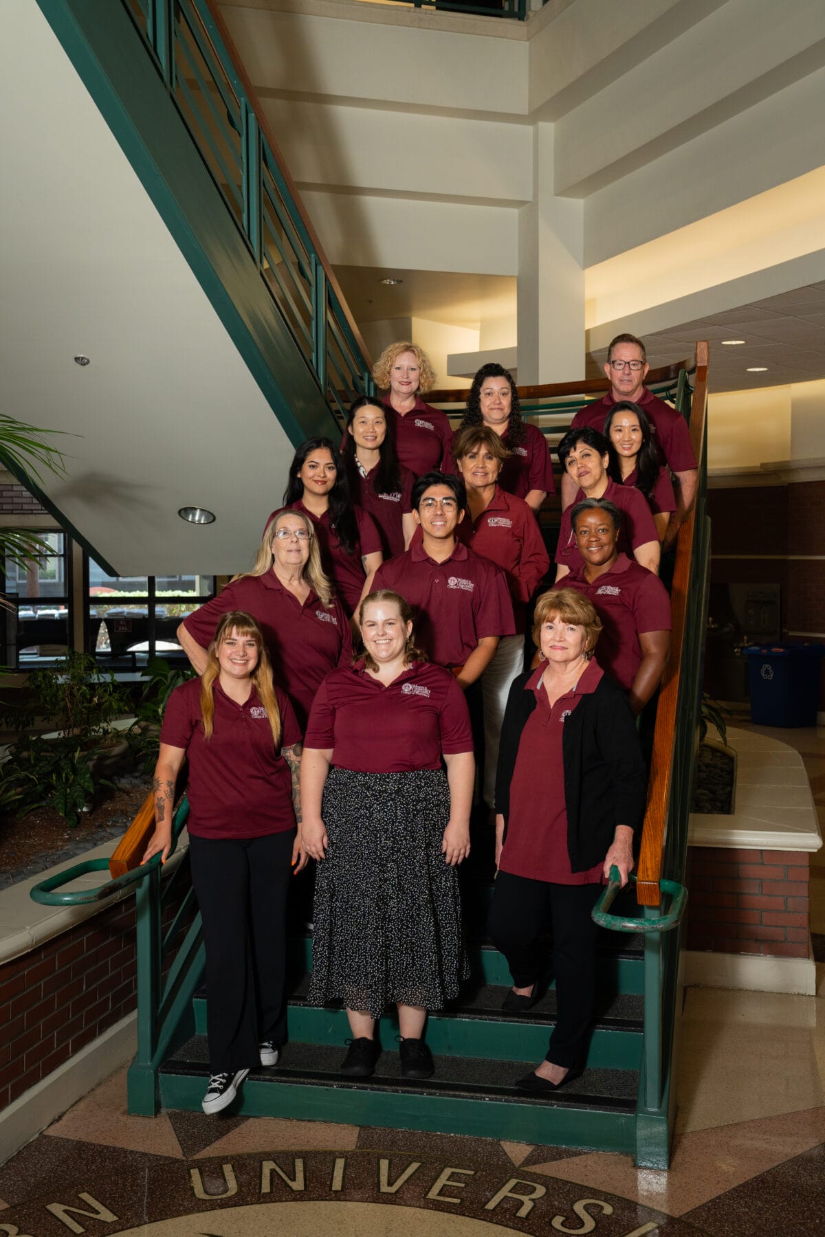 A group of fourteen people wearing maroon shirts pose on a staircase inside a building with university signage on the floor.