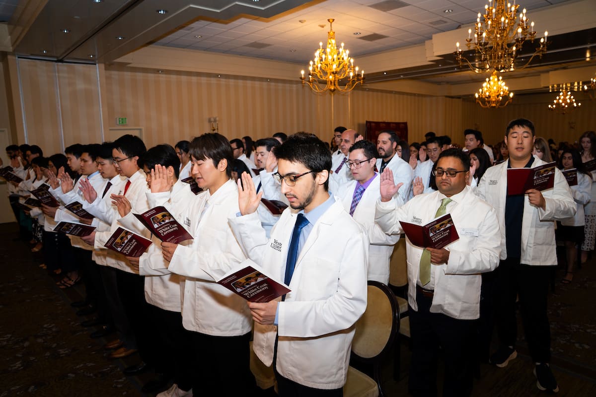 A group of people in white coats stands with right hands raised, holding booklets, participating in a ceremony in a chandelier-lit room.