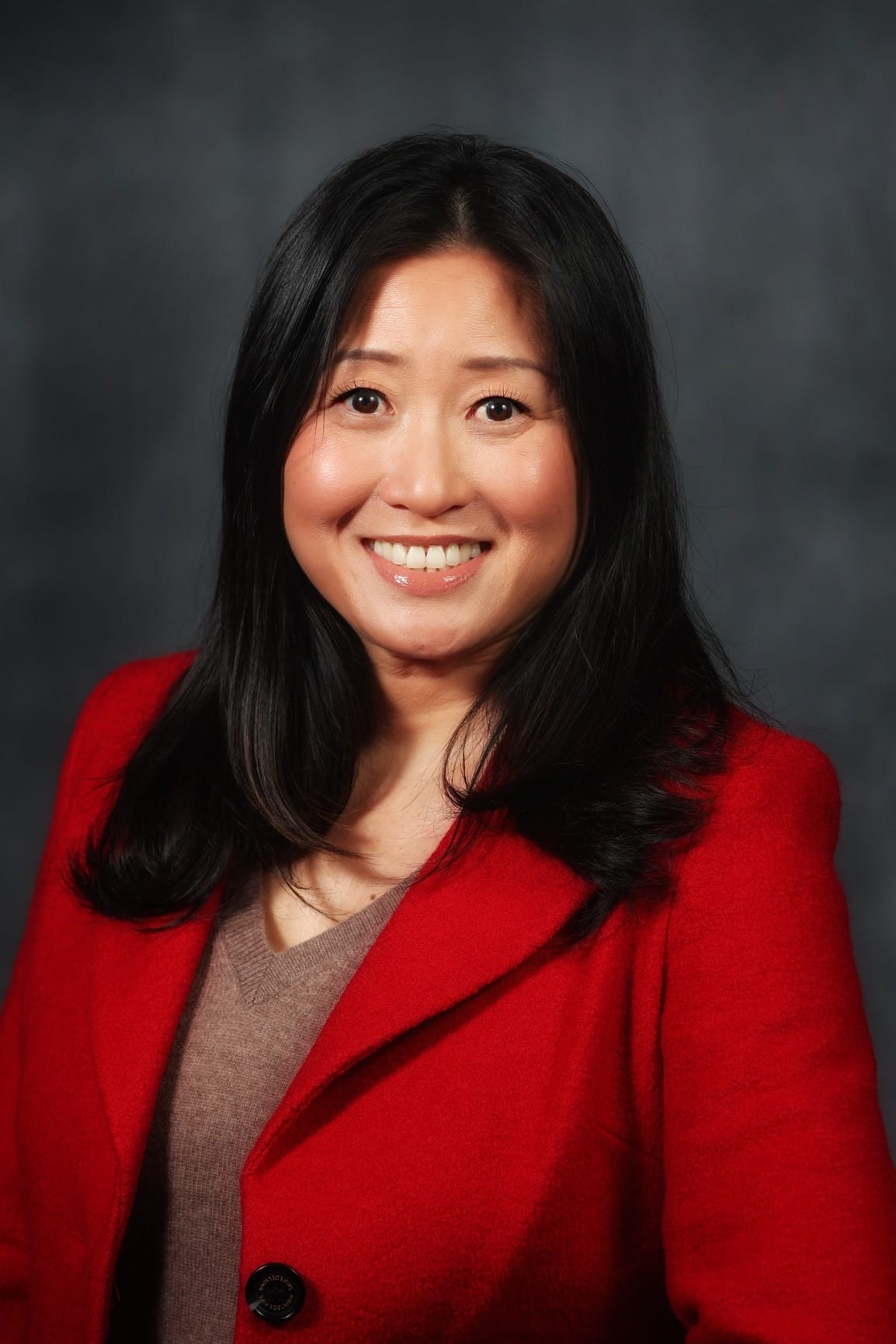 Woman with long black hair wearing a red blazer and brown top, smiling in front of a dark, blurred background.