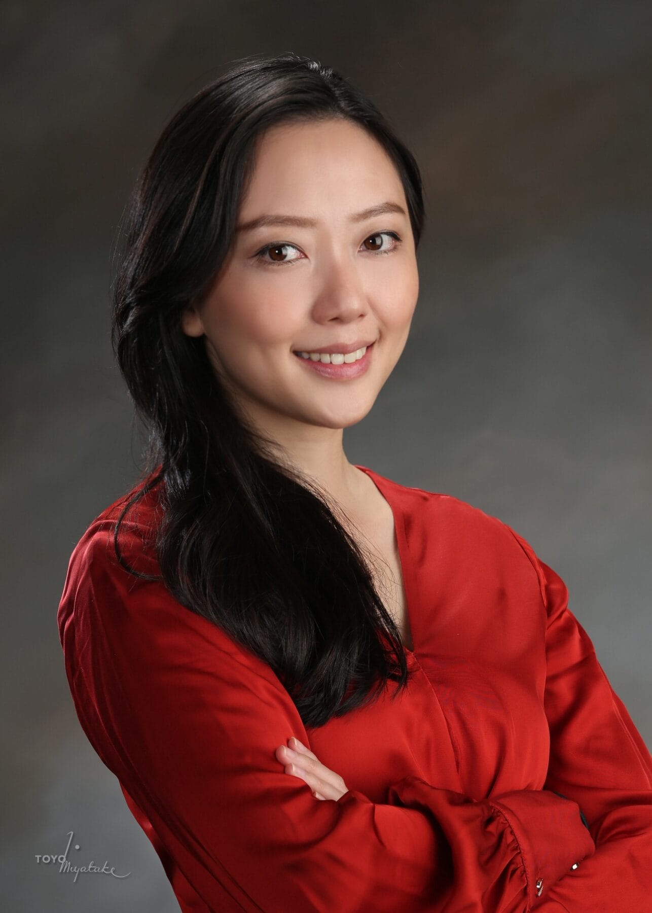 Woman with long black hair wearing a red blouse stands with arms crossed and smiles at the camera against a dark, neutral background.