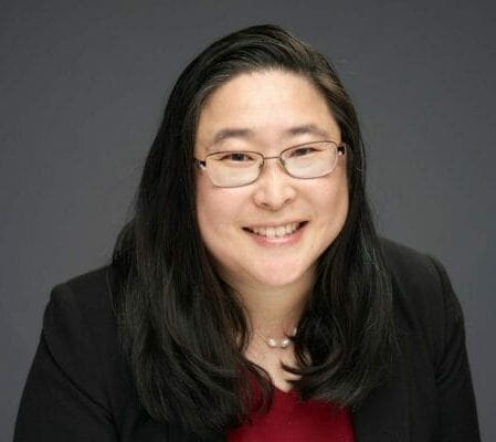 A woman with long dark hair and glasses is smiling at the camera. She is wearing a black blazer over a red top. The background is plain and dark.