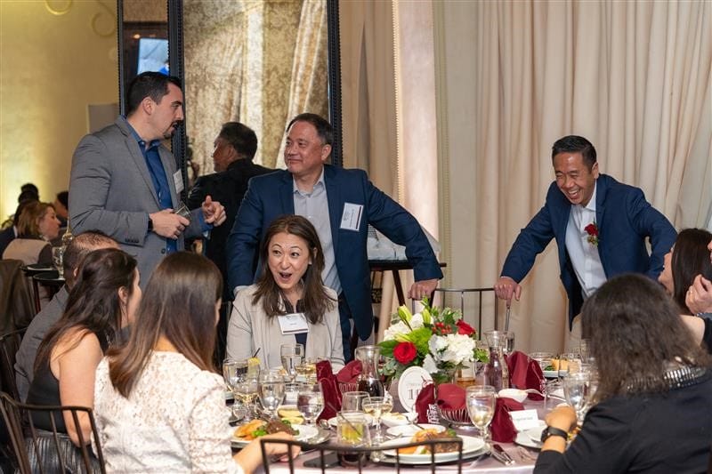 A group of people in business attire talk and laugh around a banquet table set with food, drinks, and floral centerpieces at a formal event.