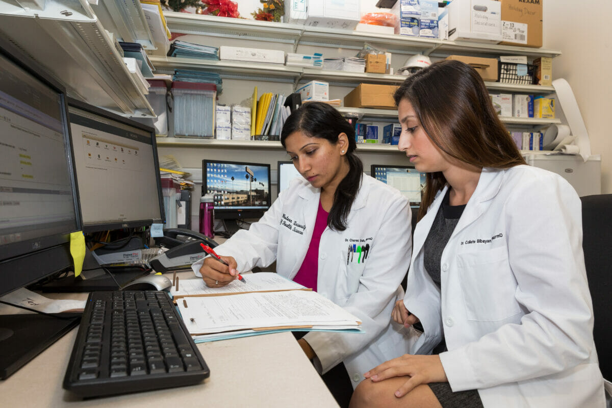 Two female students in white coats, reviewing notes, seated in front of a computer