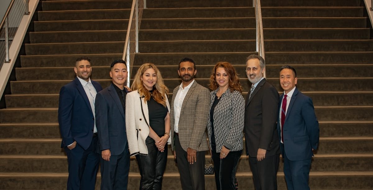 A group of seven people, dressed in business attire, stands in a line in front of a staircase and poses for a photo.