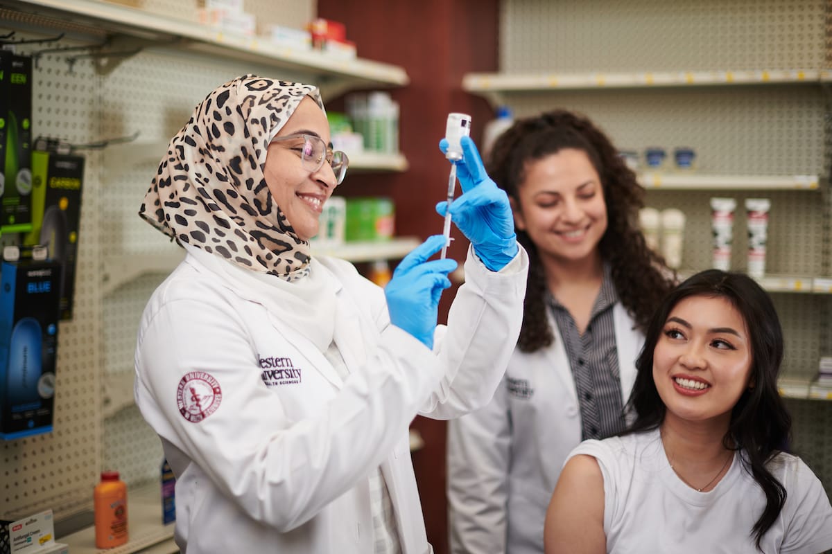 A pharmacist wearing a hijab and gloves prepares a syringe while two women look on in a pharmacy setting.