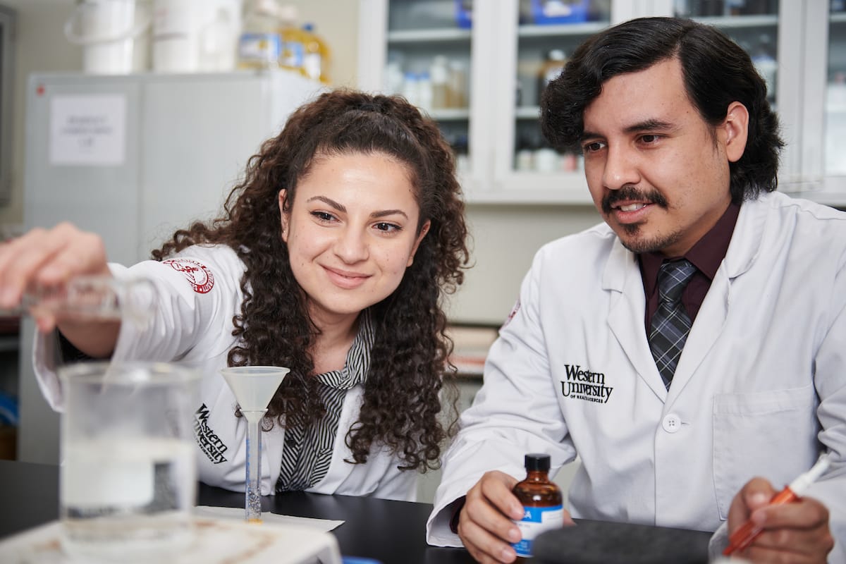 Two scientists in lab coats conduct an experiment, pouring liquid into a beaker on a lab table, with scientific equipment and shelves in the background.
