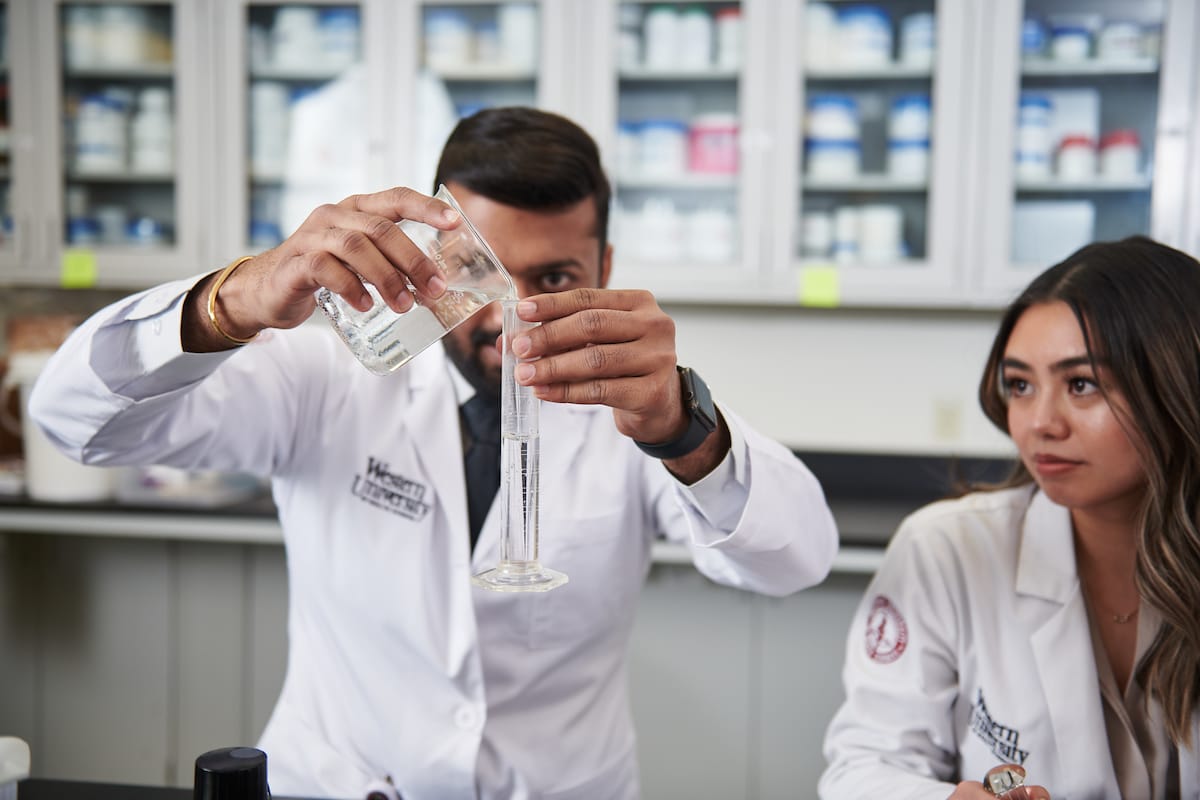Two individuals in white lab coats are in a laboratory. One is pouring liquid from a beaker into a graduated cylinder, while the other observes. Shelves with various containers are visible in the background.