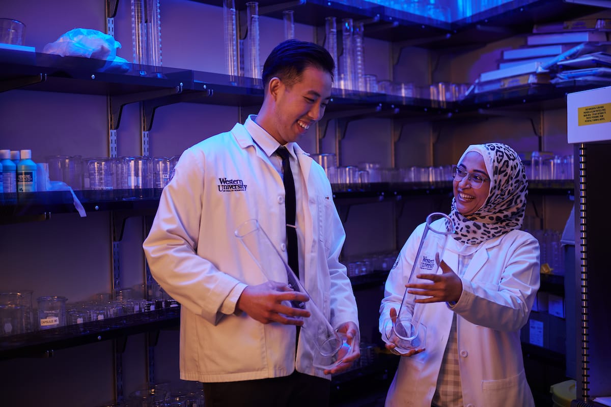 Two laboratory researchers in white coats, one wearing a hijab, are smiling and examining lab glassware in a blue-lit lab. Shelves with various bottles and containers are visible in the background.