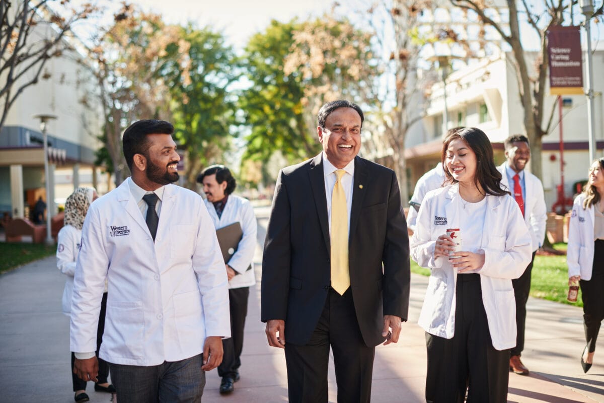 A group of people, including men and women in white lab coats, walk together outdoors, led by a man in a dark suit and yellow tie, on a sunny day. Trees and buildings are visible in the background.