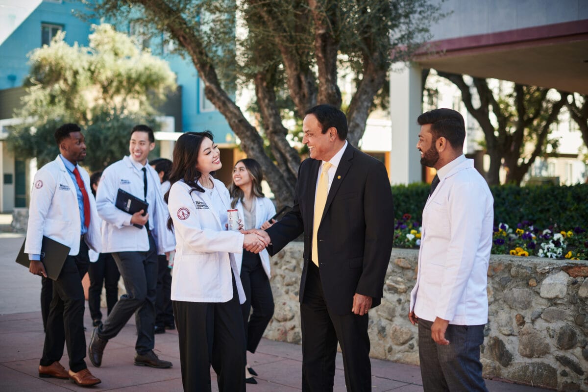 A group of medical professionals, wearing white coats, engage in conversation and handshake with a man in a black suit outdoors. Several people stand in the background.