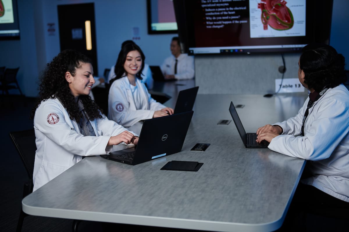 Three medical students in white lab coats work on laptops at a conference table, with anatomical diagrams displayed on screens in the background.