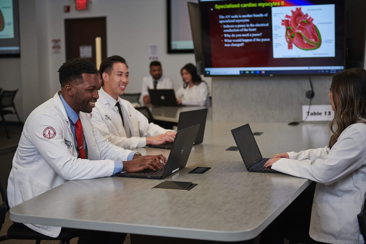 Four medical students in white coats sit around a table with laptops, engaging in a study session. A presentation on cardiac myocytes is displayed on a screen in the background.