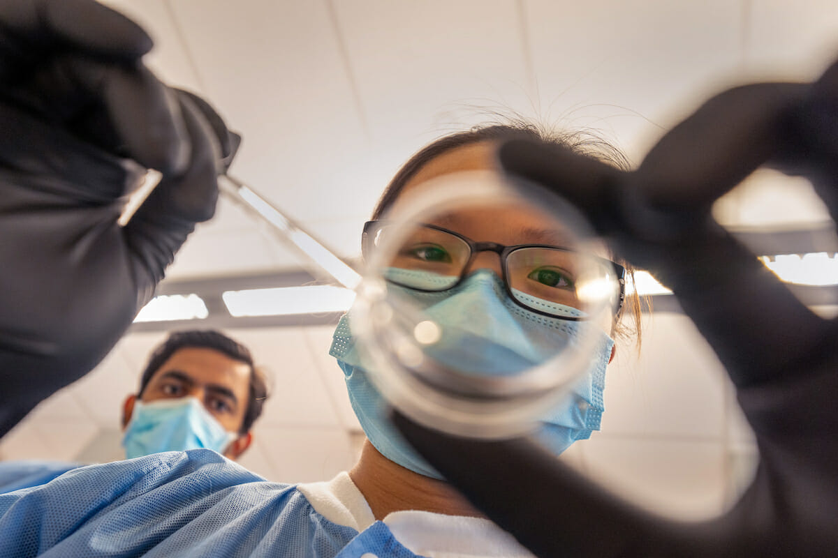 Two people wearing surgical masks and gloves looking at a glass.