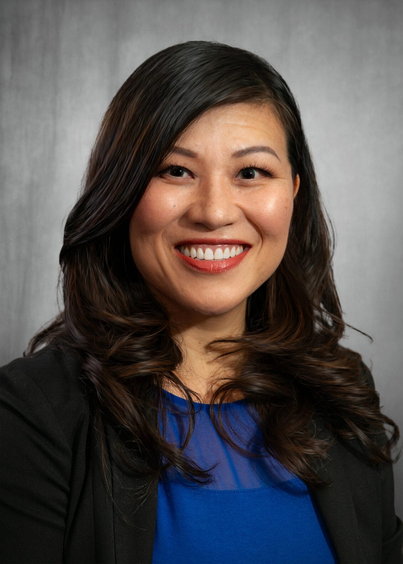Woman with long dark hair in a black blazer and blue top smiles at the camera in front of a gray background.