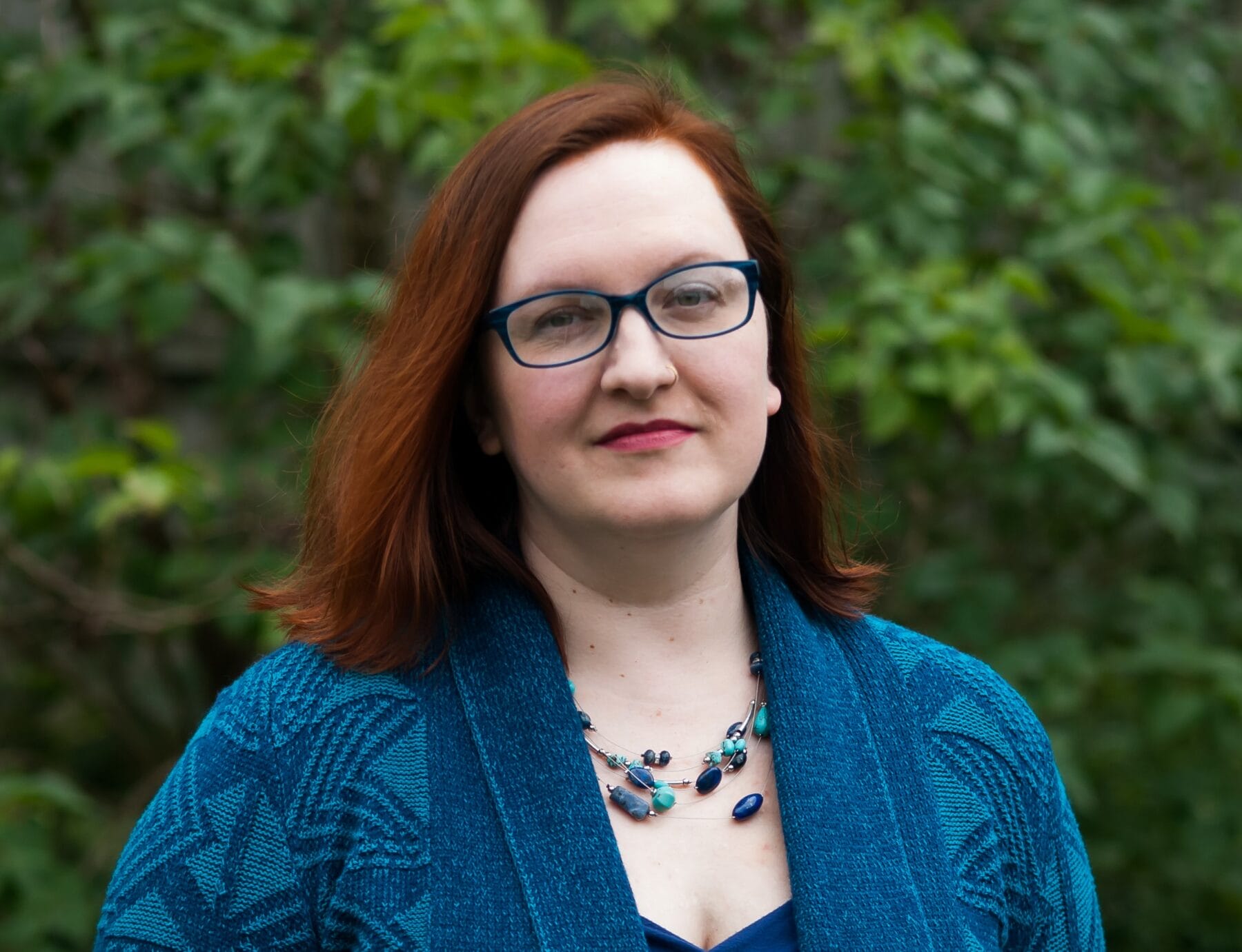 Person with red hair and glasses wearing a blue sweater and necklace, standing outdoors with green foliage in the background.