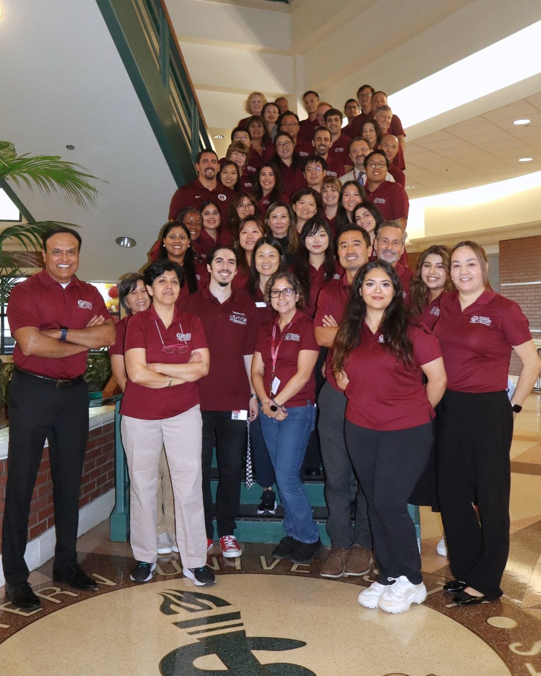 A large group of people wearing matching maroon shirts poses together on a staircase inside a building with natural light.
