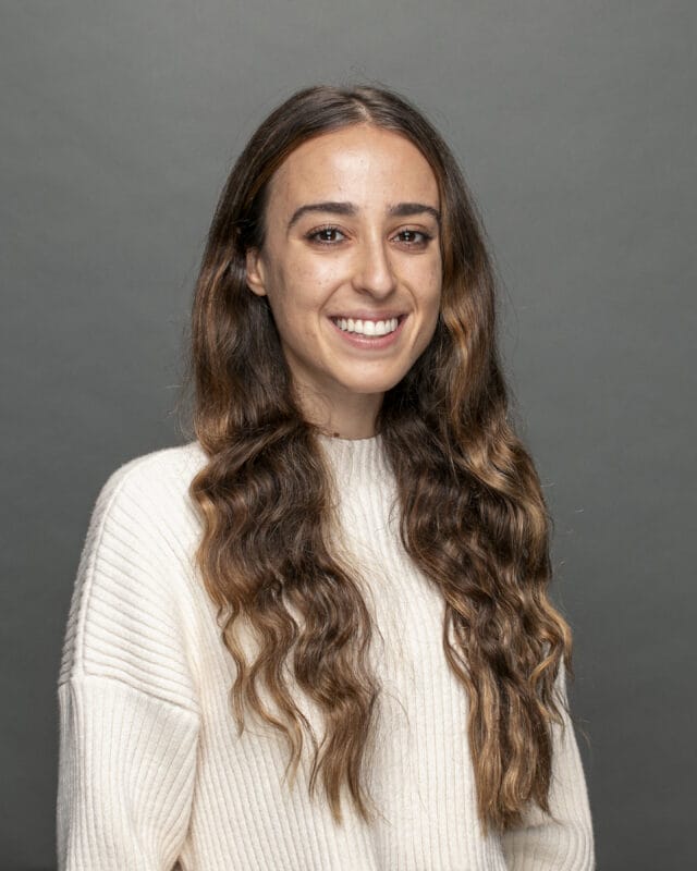 A woman with long, wavy brown hair and a white sweater smiles against a gray background.