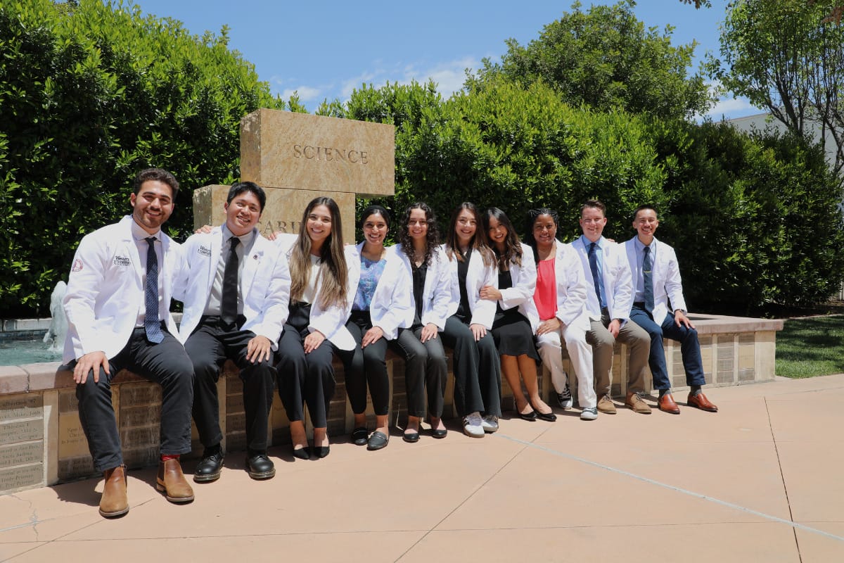 A group of ten people, all wearing white lab coats, pose for a photo while sitting on a stone wall outdoors in front of a building sign reading "Science." Trees and shrubs are in the background.