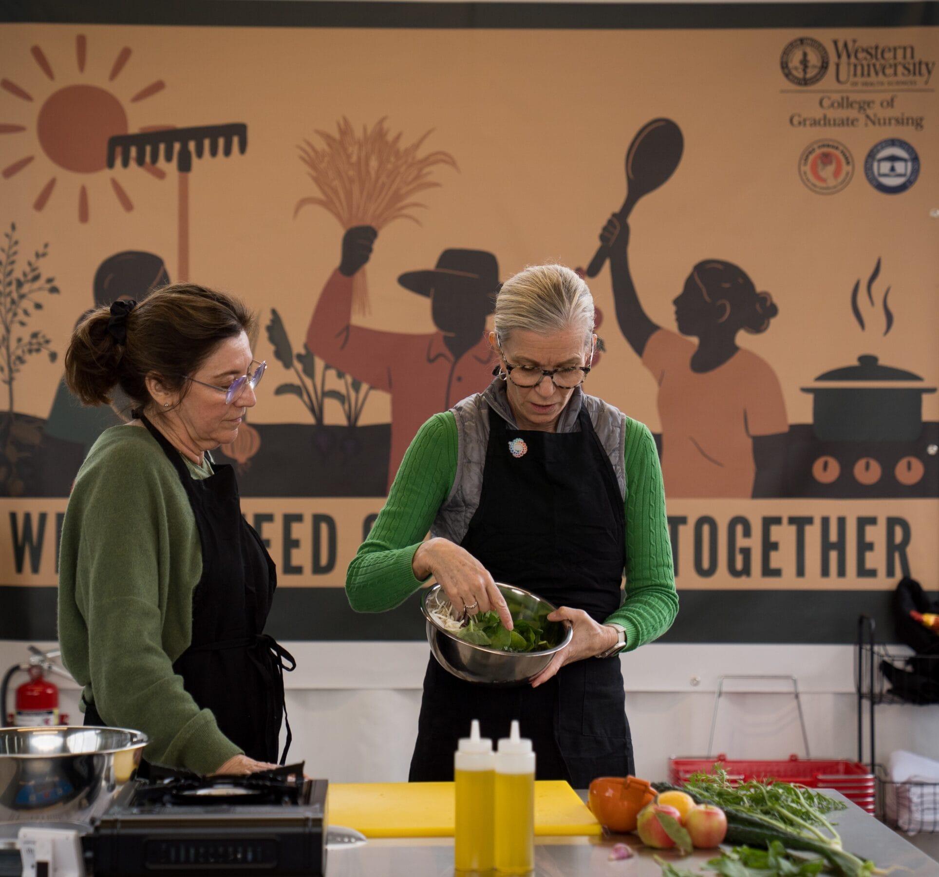 Two women wearing aprons prepare food together in front of a mural and table with vegetables, bottles, and cooking utensils at a Western University College of Graduate Nursing event.