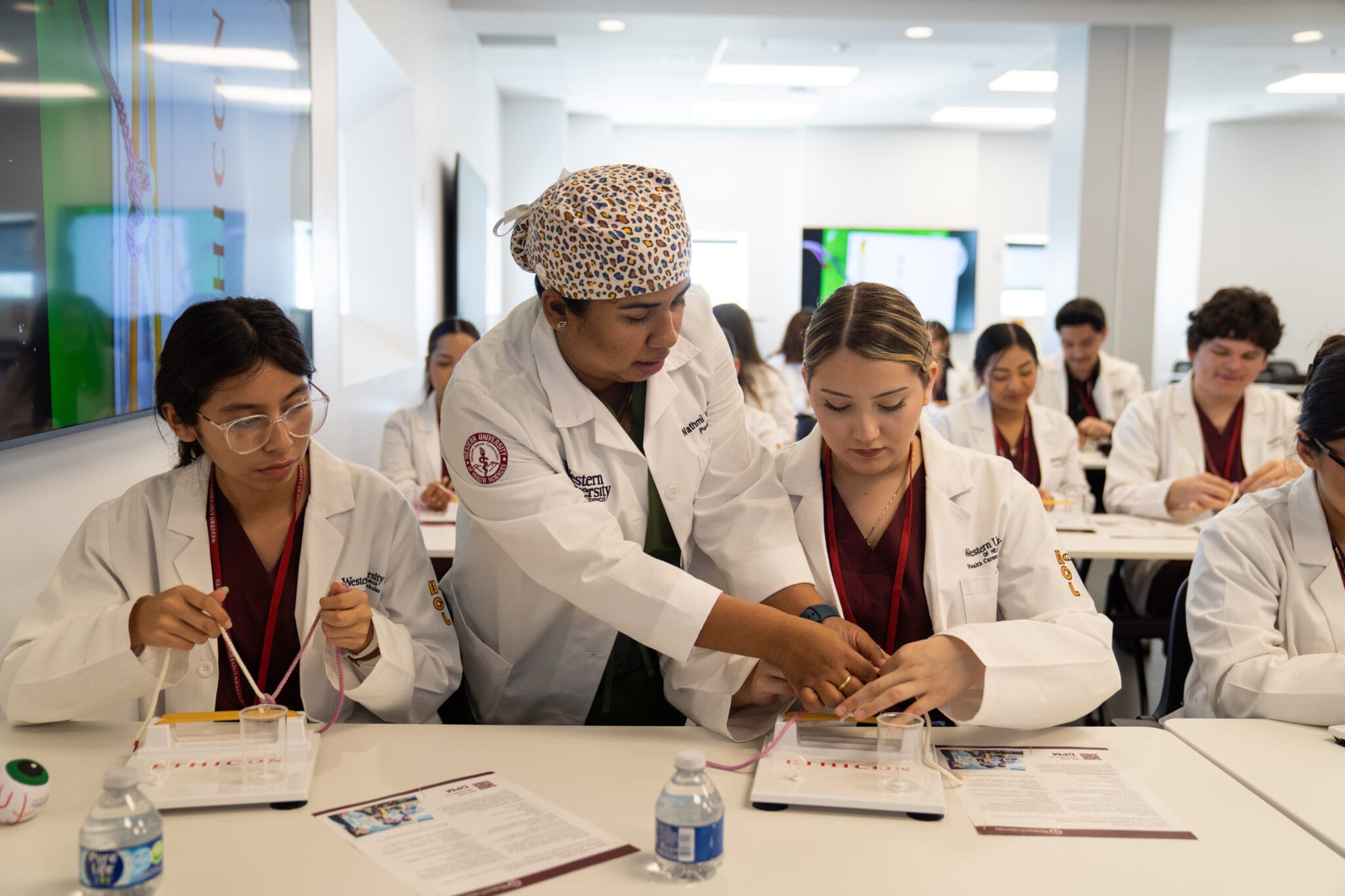 A medical instructor demonstrates a procedure to students in lab coats working at a classroom table with supplies, notebooks, and water bottles.