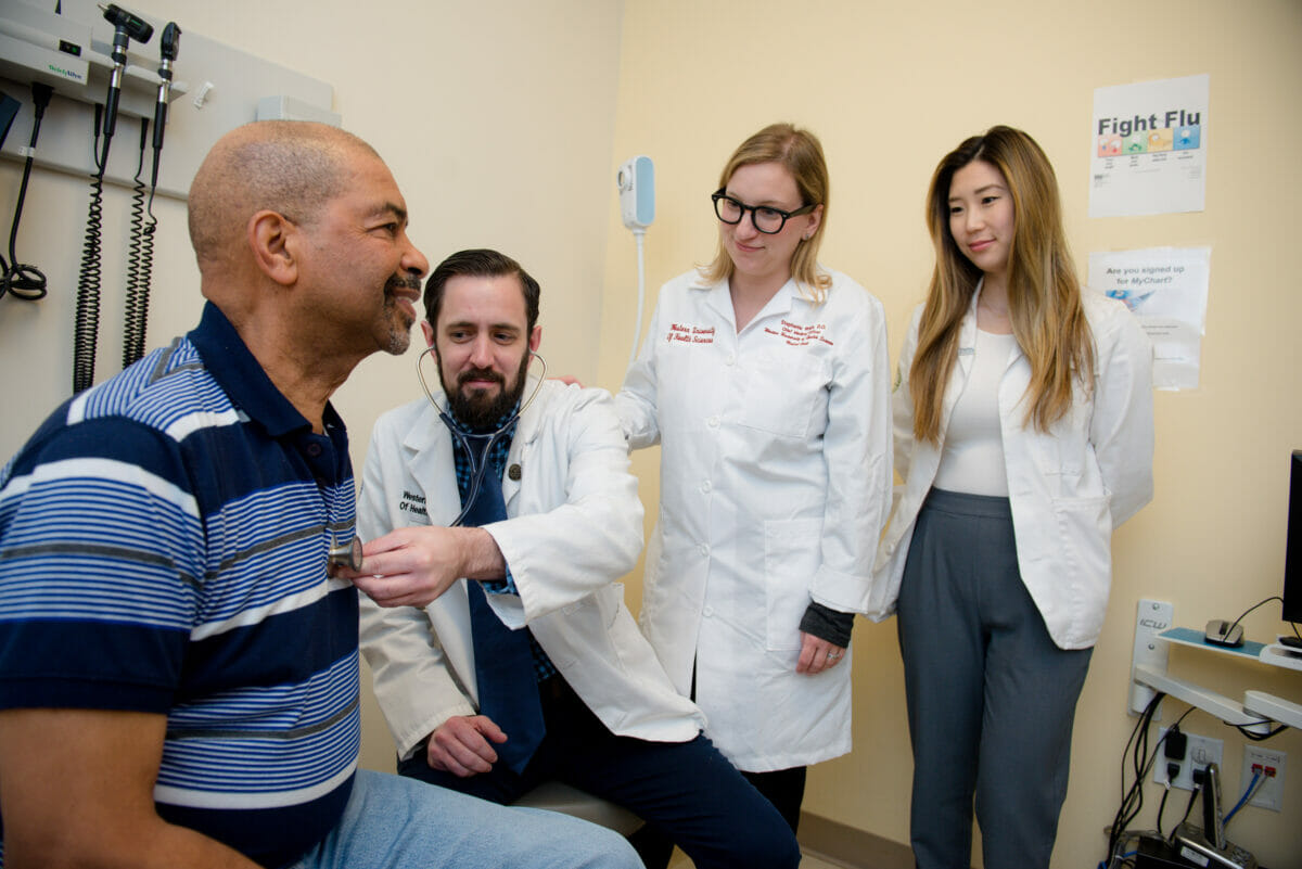 Image of a PA student examining a patient. Being observed by a COMP doctor and a COMP student