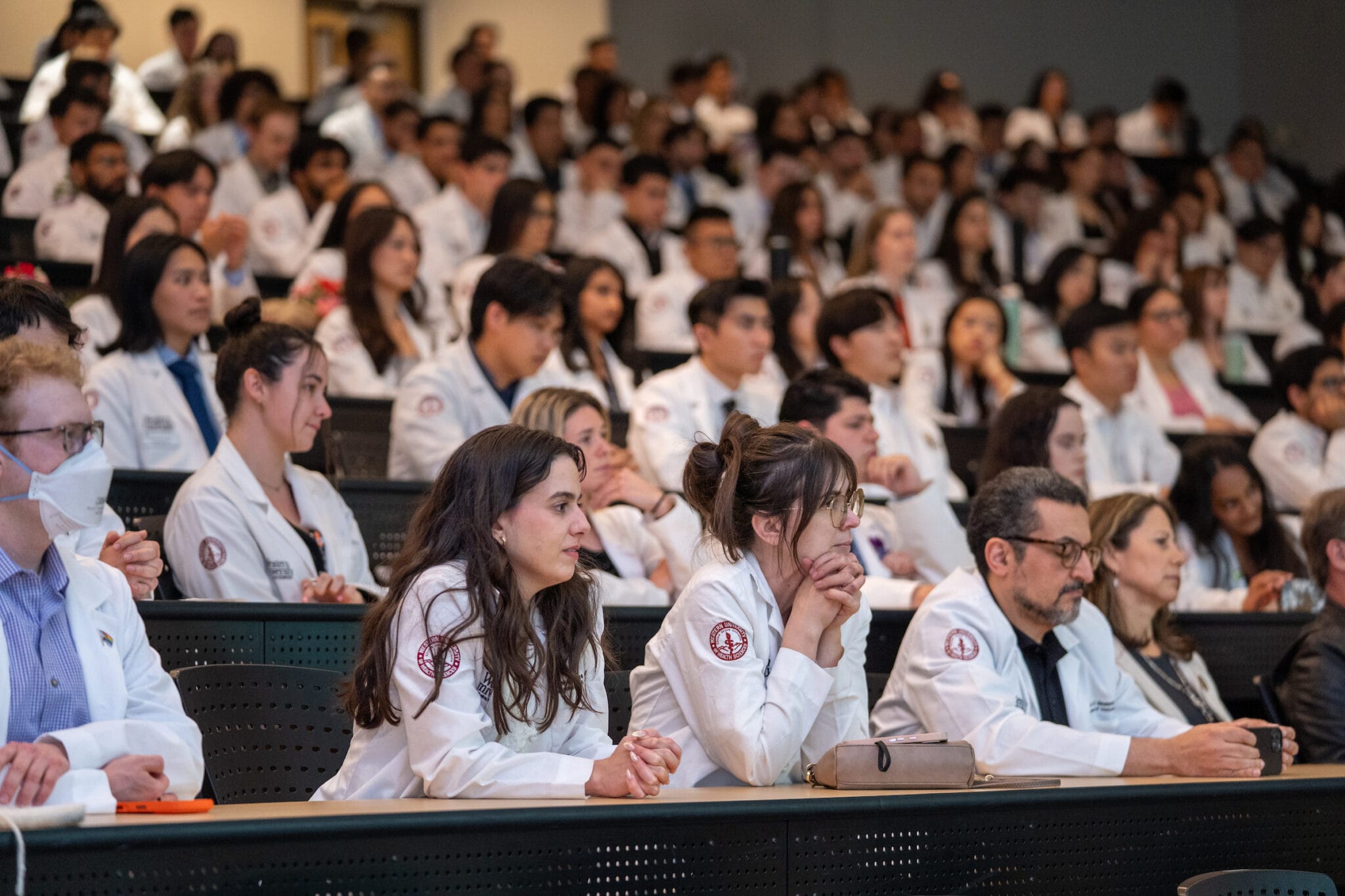 A large group of medical students and professionals in white coats sit attentively in an auditorium during a lecture or presentation.