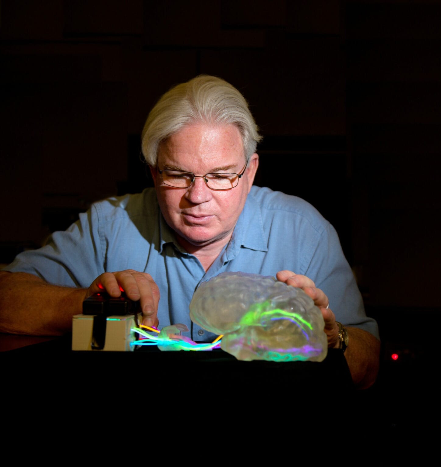 An older man in glasses examines a translucent brain model with colorful lights, adjusting wires connected to a small electronic device on a dark table.