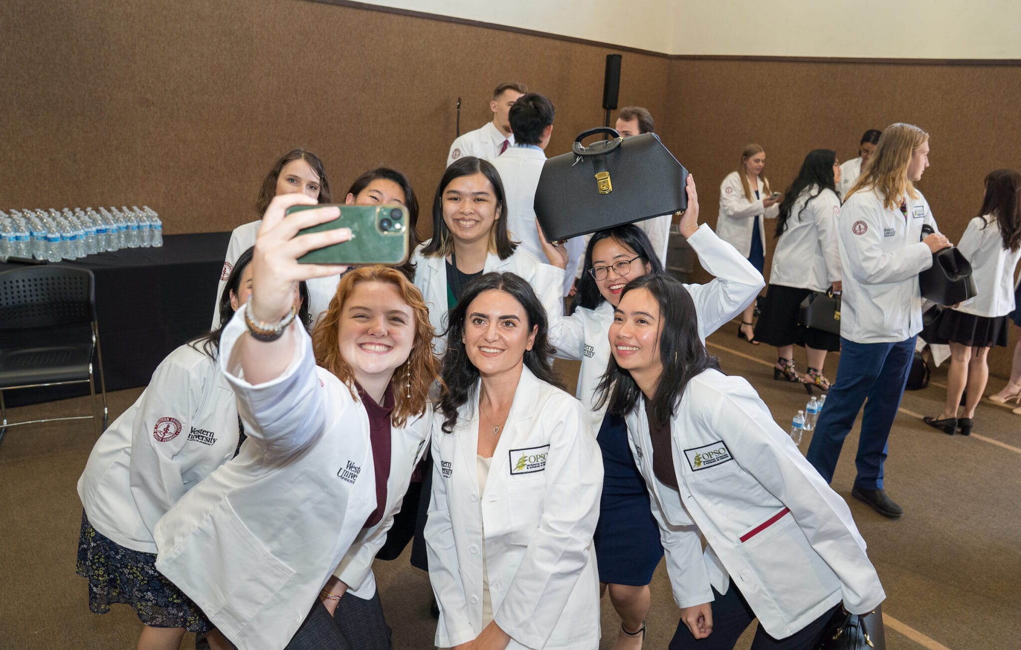 A group of students in white coats take a selfie together indoors at an event while others stand and talk in the background.