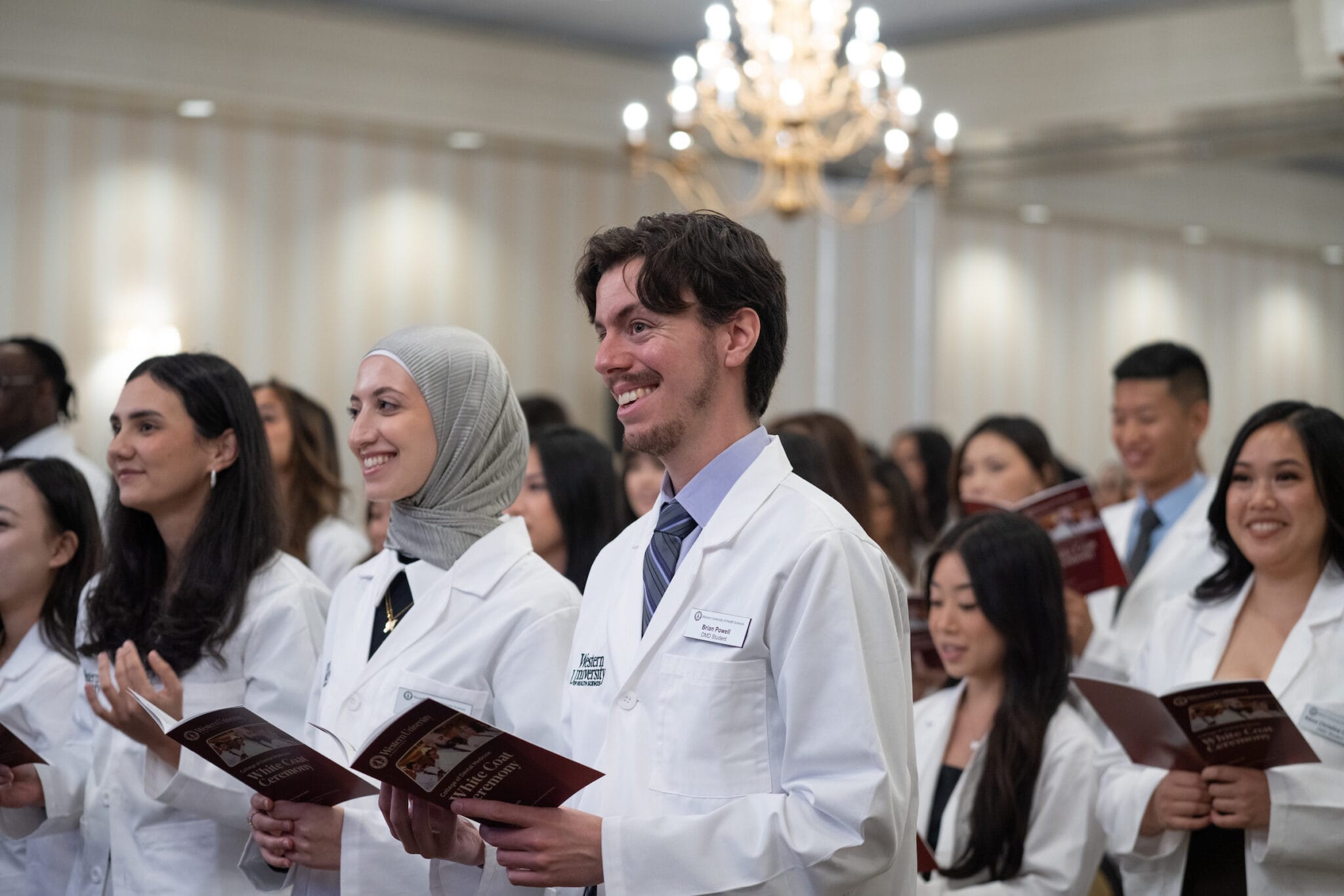 A group of medical students in white coats hold programs and smile during a ceremony in a formal, well-lit room with a chandelier.