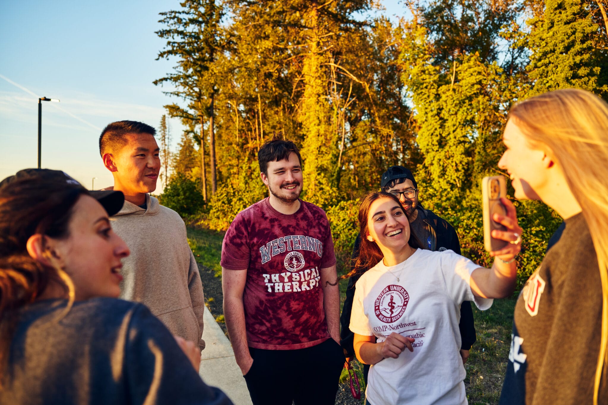 A group of young adults standing outside, some smiling and laughing, while one person holds up a phone for a selfie. Trees are visible in the background.