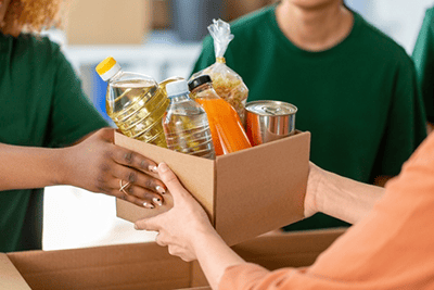 A person hands a box of assorted food items to another person. The box contains bottled oil, water, juice, pasta, and a canned good.