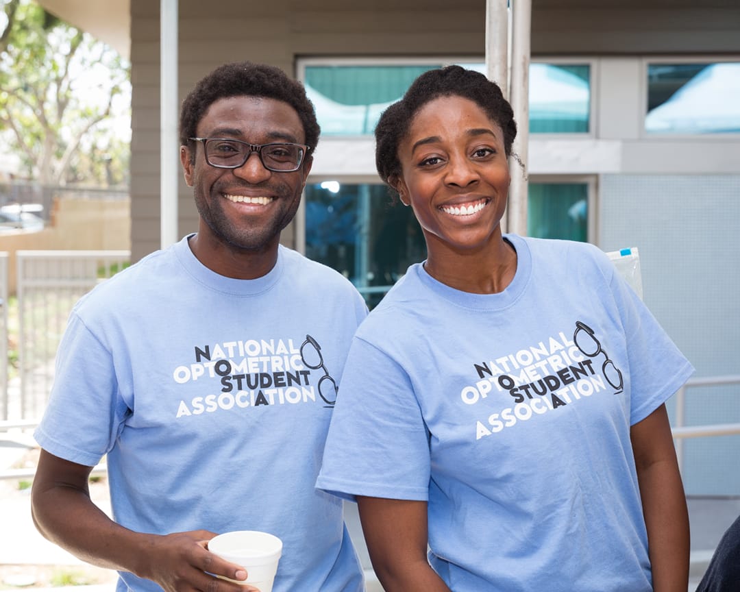 Two people wearing matching "National Optometric Student Association" t-shirts smile at the camera outdoors. One holds a white cup.
