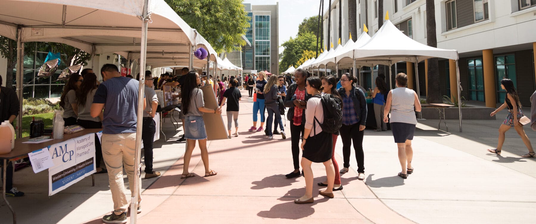 People walk and gather at outdoor booths under tents along a sidewalk between campus buildings on a sunny day.