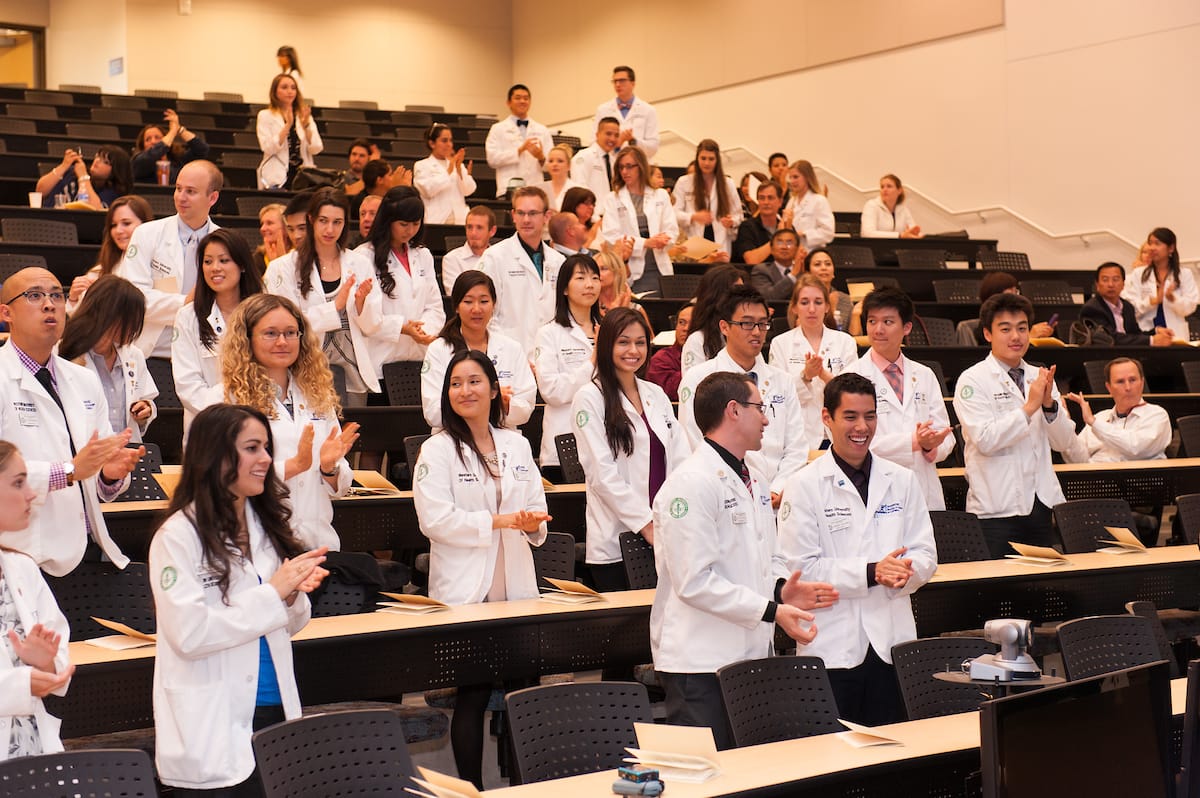 A large group of medical students in white coats stand and applaud in a lecture hall.