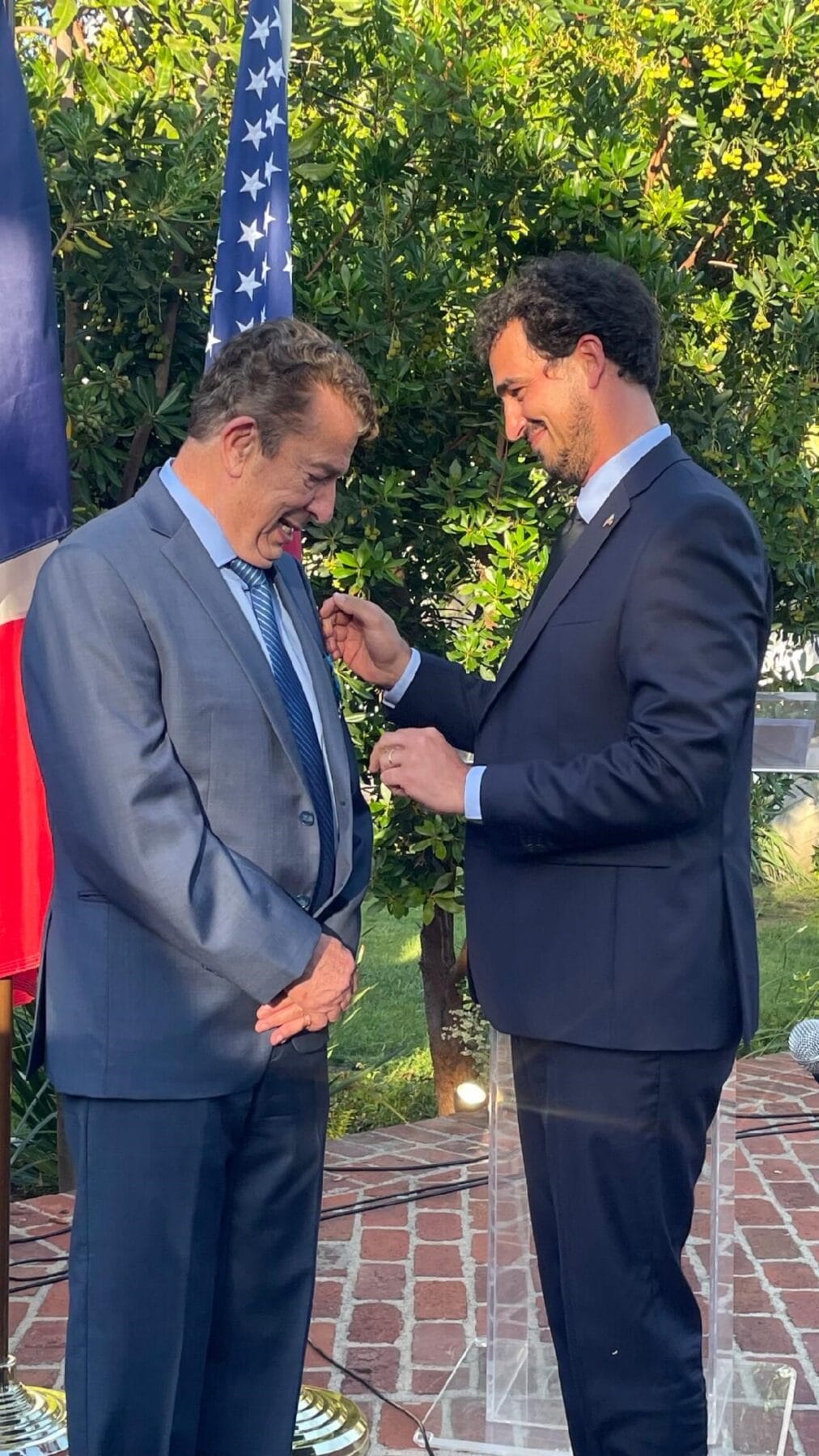 Two men in suits stand outdoors; one man pins a medal onto the other’s lapel, with flags and greenery in the background.