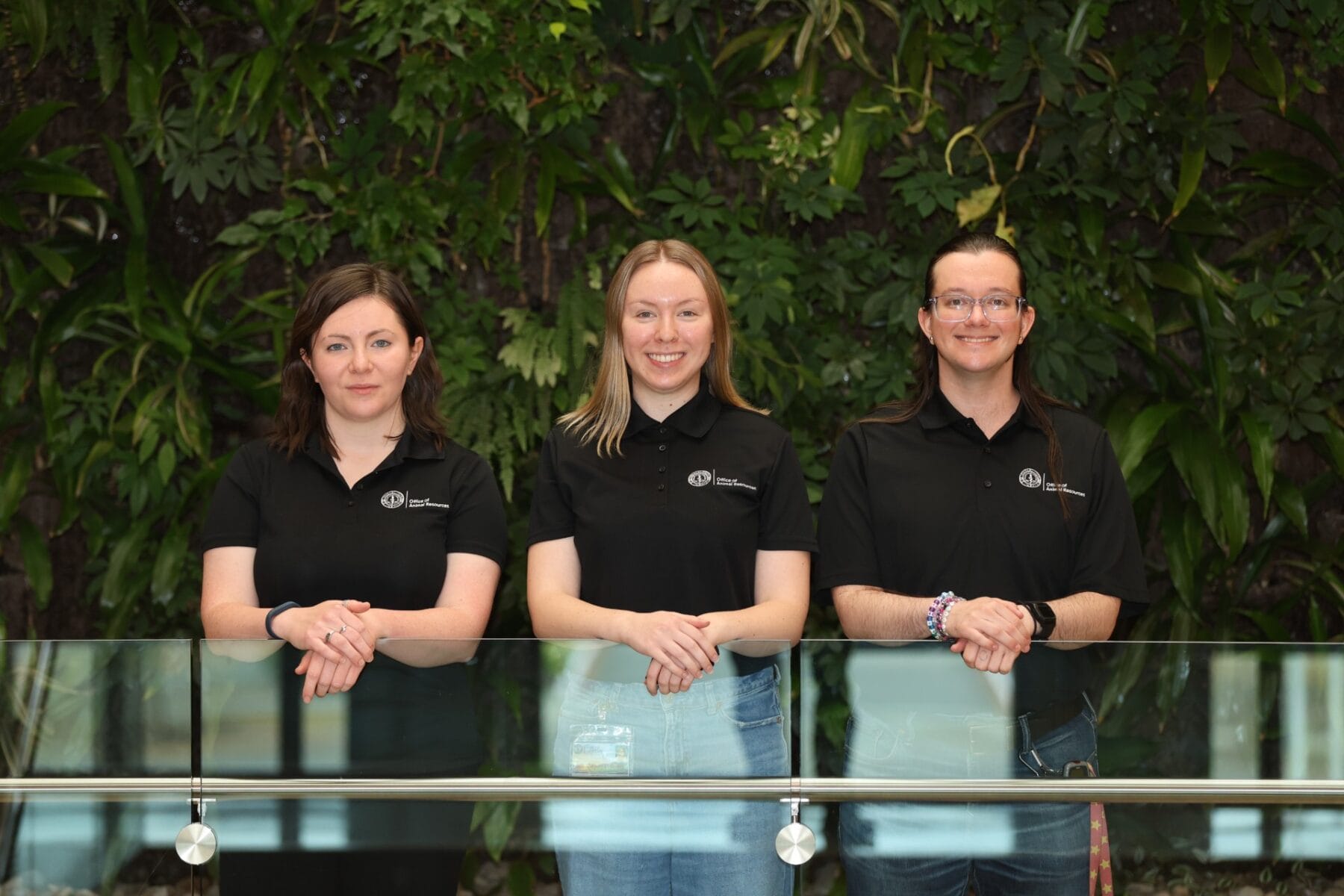 Three people wearing black polo shirts stand side by side, smiling, in front of a green leafy background and a glass railing.
