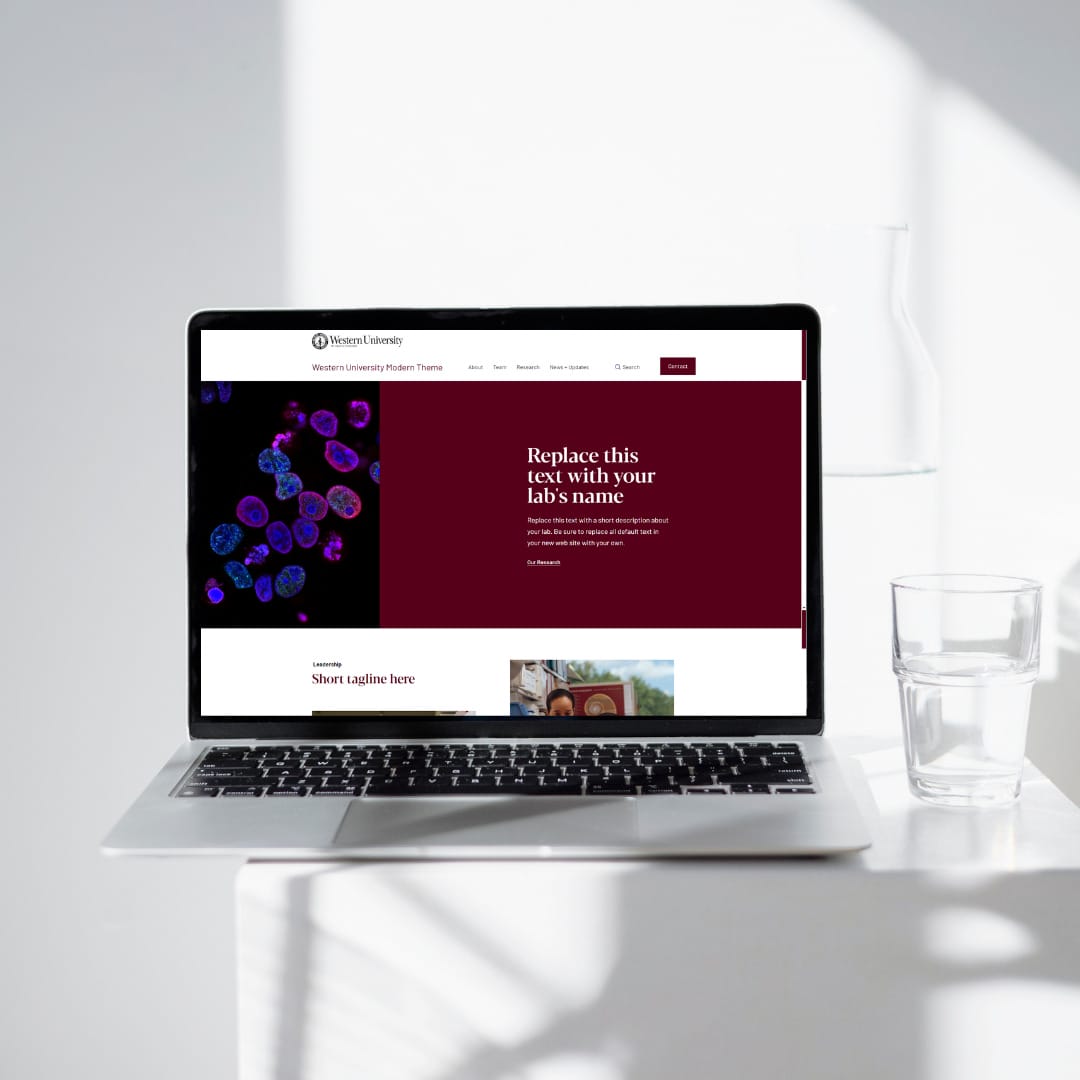 A laptop on a table displaying a website template for a research laboratory. There is a glass of water next to the laptop.