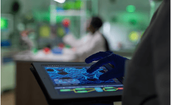 A person in gloves uses a tablet displaying scientific data in a lab, with another person working in the blurred background.