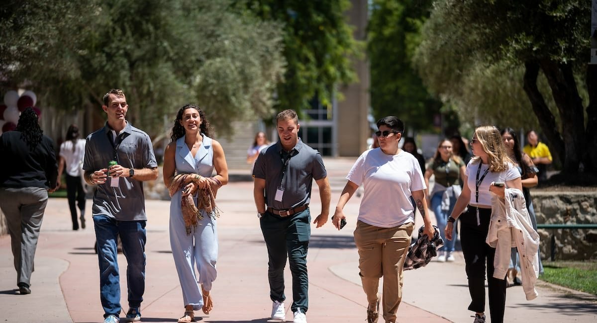 A group of people walk along a sunlit pathway lined with trees, casually conversing and smiling, others walk in the background.