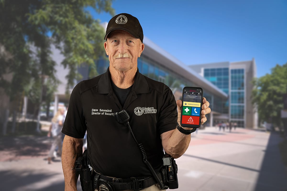 A security officer stands outside a building, holding a smartphone displaying an emergency app. He's dressed in uniform with a radio and badge visible. The background shows a sunny day on a modern campus.