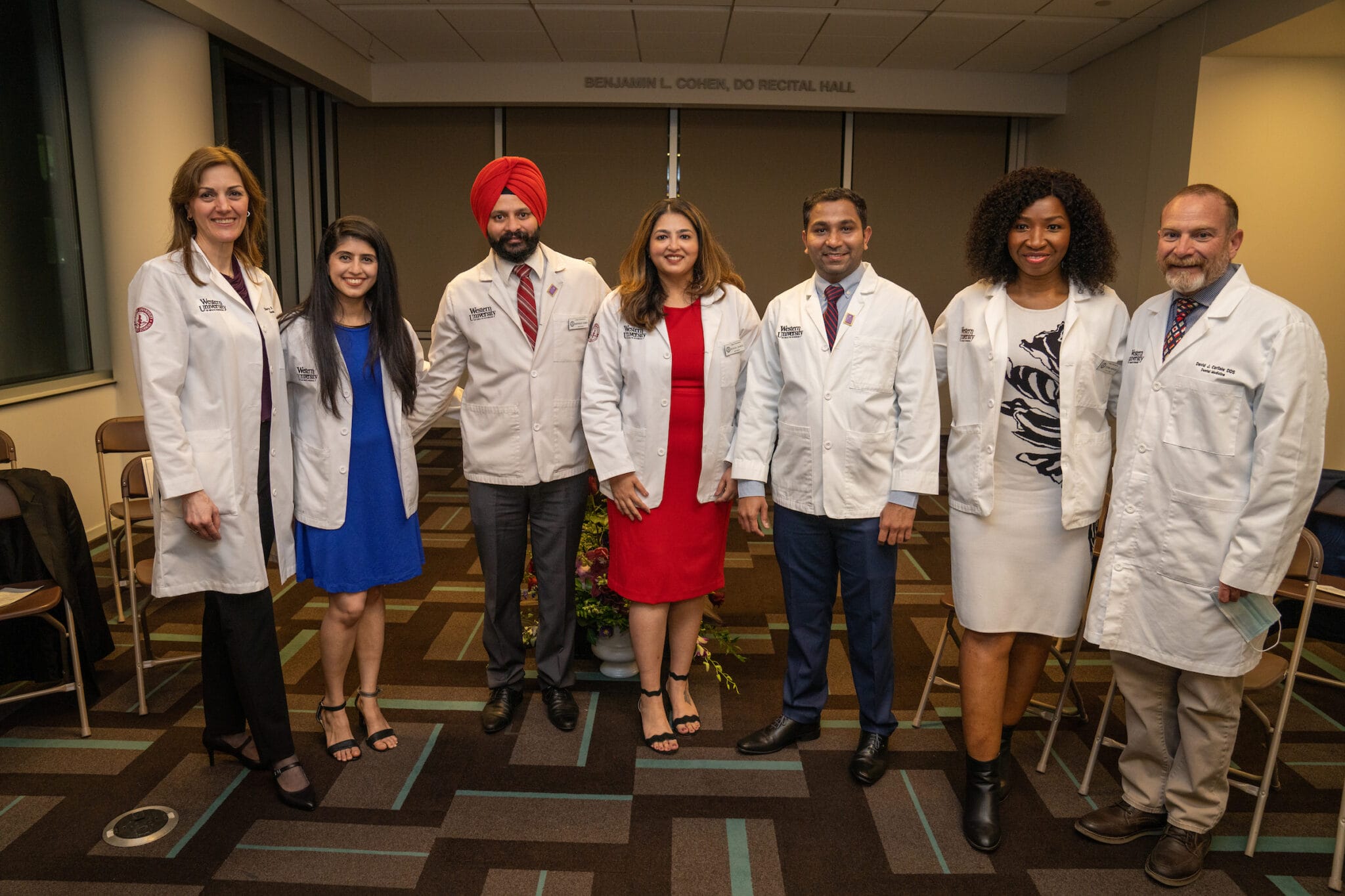 Seven people in white lab coats stand indoors in a row, smiling for a group photo in a professional setting.