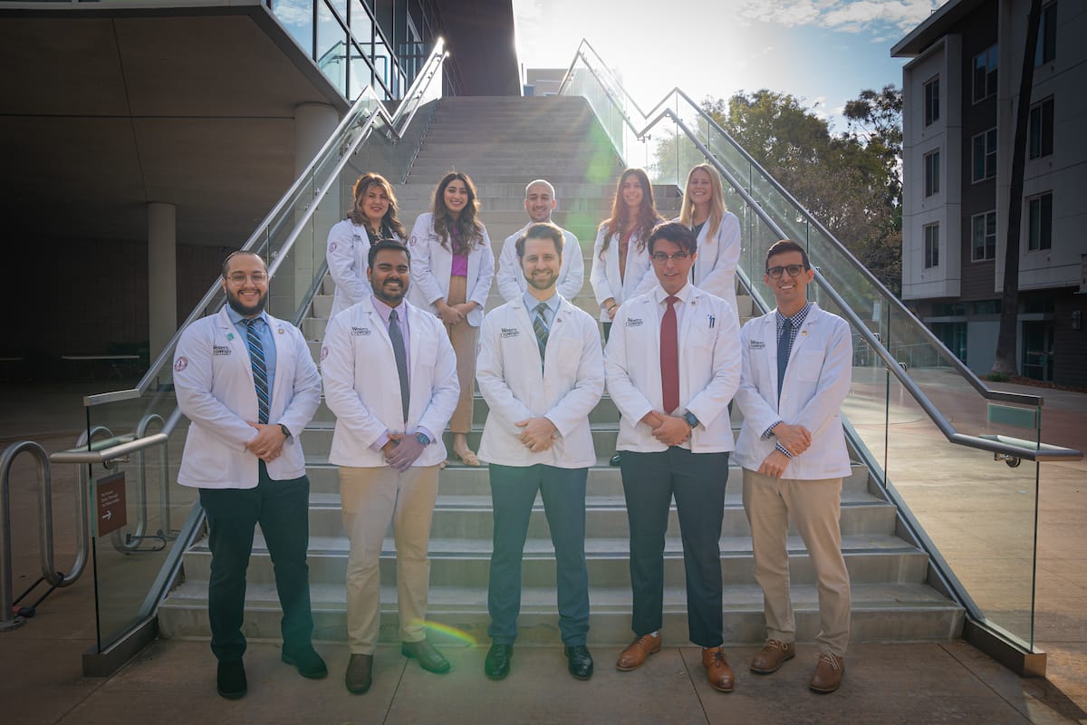 A group of ten people wearing white coats stands in two rows on an outdoor staircase. The sun shines brightly behind the stairs.