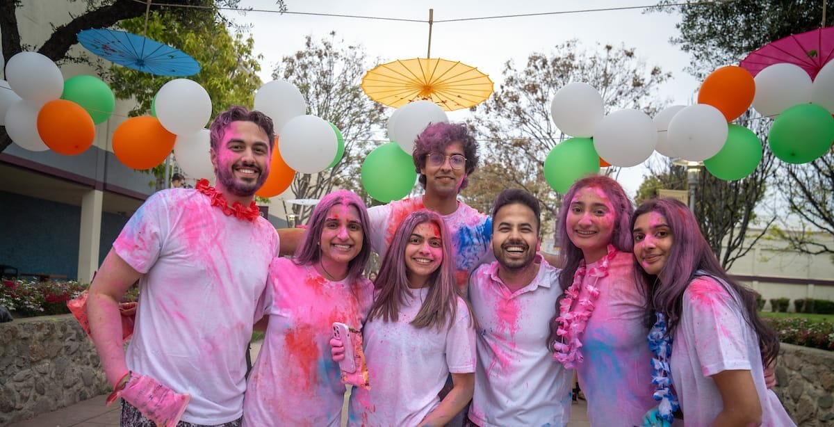 A group of people covered in colorful powder stands together, smiling and posing for a photo under a decorative setup with balloons and umbrellas.