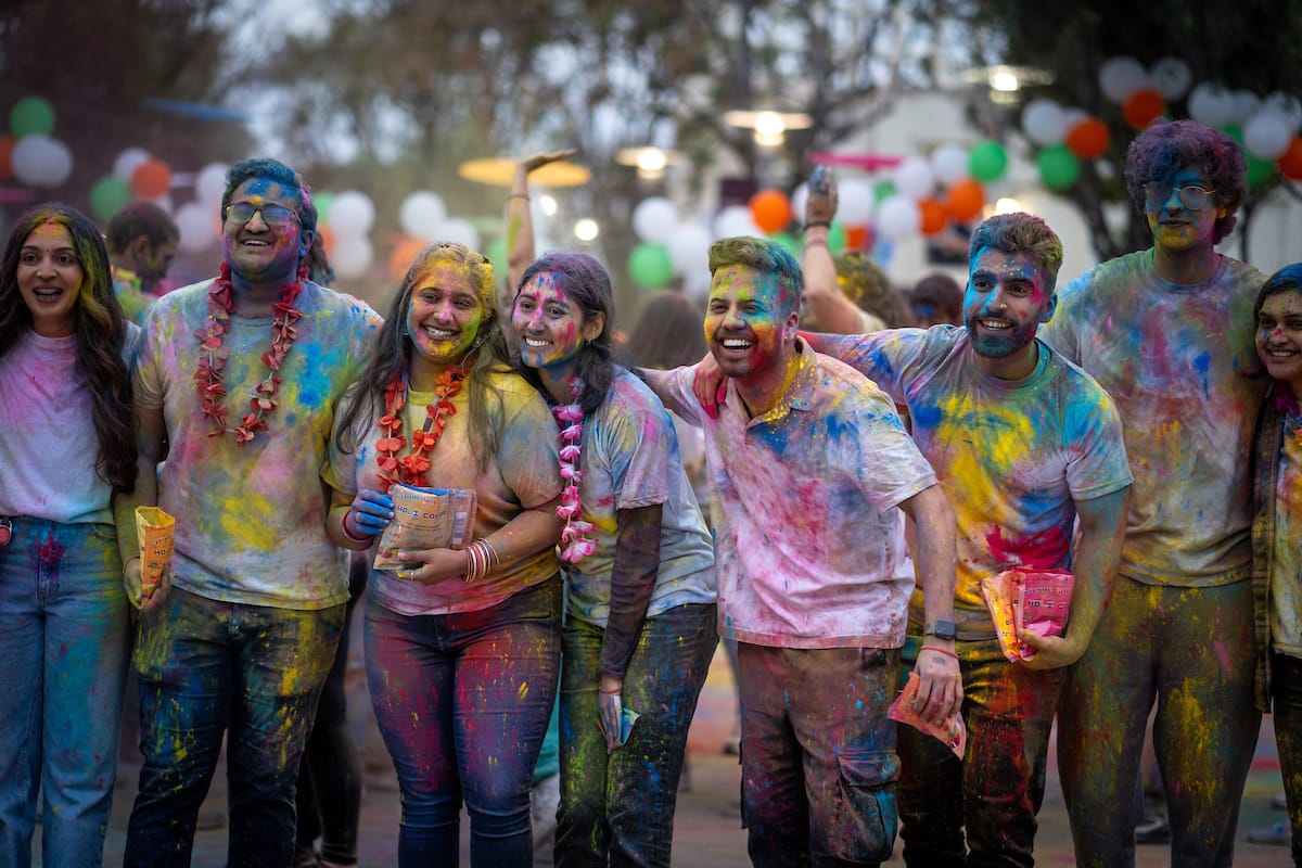 A group of people covered in vibrant colored powders stand together, smiling and posing for a photo during a festive outdoor event. Some are holding small bags with remaining powder.