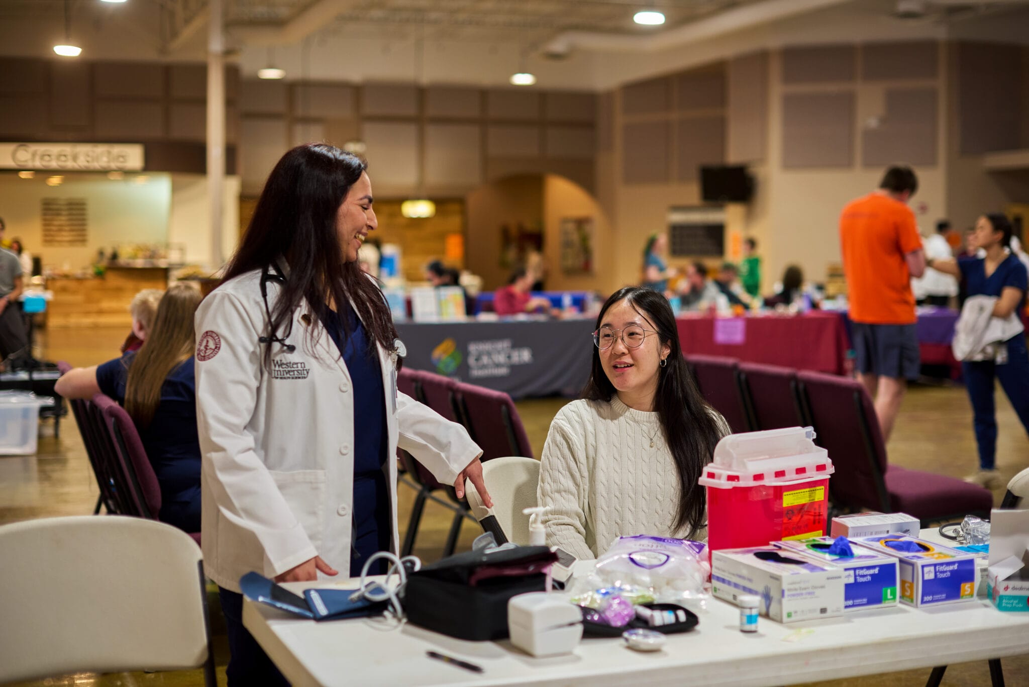 A healthcare worker in a white coat stands next to a seated woman at a table with medical supplies in a large indoor clinic setting.