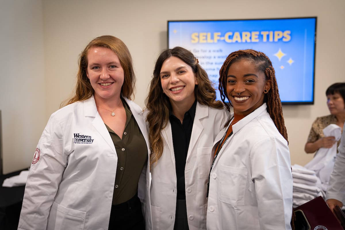 Three women in white lab coats smile and stand together. In the background, a screen displays "Self-Care Tips.