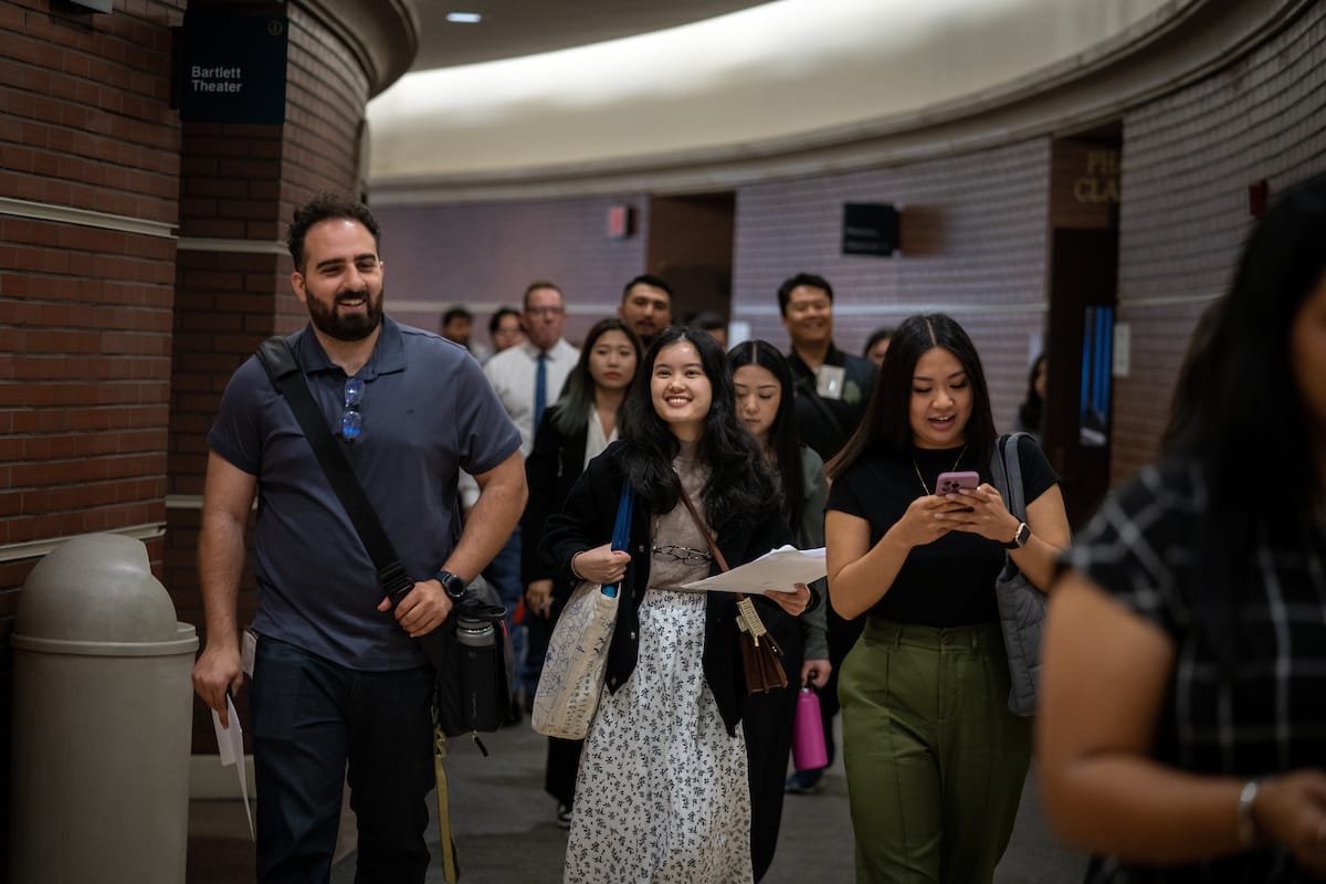 A group of people walking in a hallway. The man on the left is carrying a bag, the woman in the center is holding papers, and the woman on the right is looking at her phone.